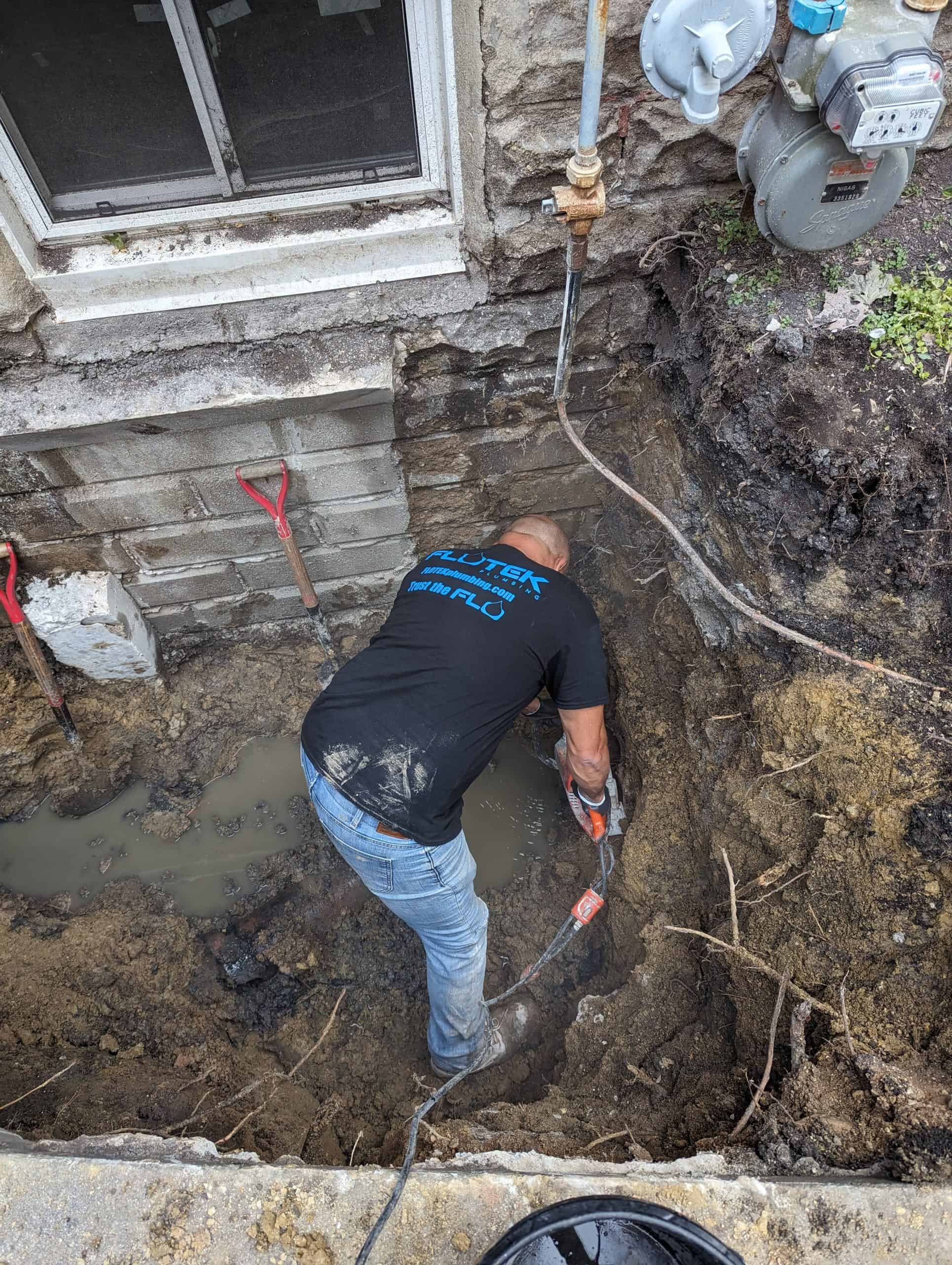 A person is digging in a muddy area next to a building's foundation. They are using a power tool and wearing a black shirt and jeans. Red shovels are visible nearby, and water is pooled in the trench. There's a gas meter on the wall above.