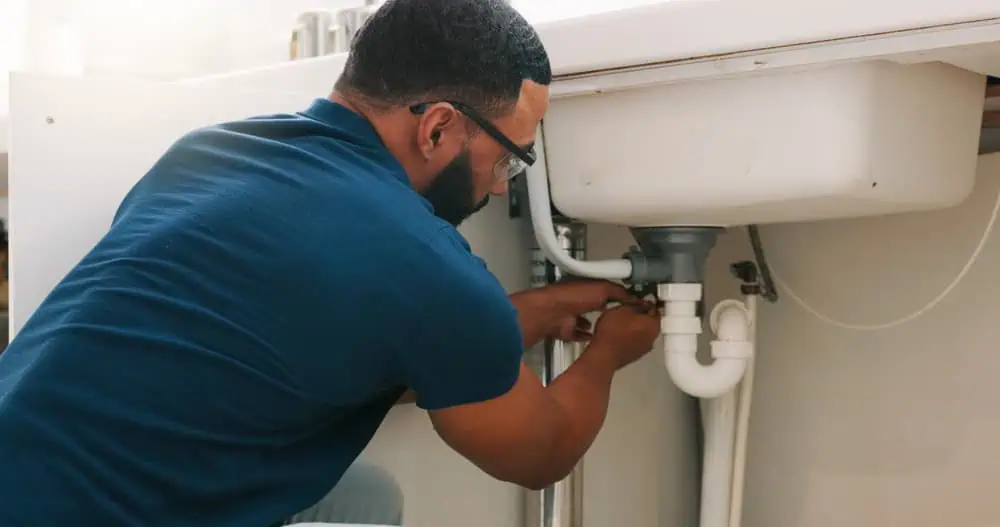 A Black professional plumber performing maintenance on a kitchen sink. The plumber is focused on repairing or inspecting the sink plumbing, using tools such as a wrench, pliers, and a pipe wrench. The modern kitchen features polished countertops, a stainless steel sink, and organized cabinets. A bucket is placed underneath the pipes to catch water, emphasizing careful and professional plumbing work. The image highlights expertise, precision, and hands-on plumbing service in a residential setting.