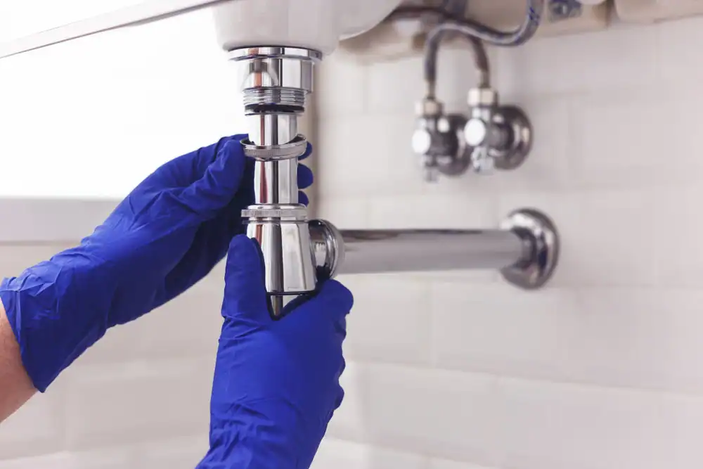 A licensed plumber in Kane County, Illinois, is kneeling beneath a bathroom sink while repairing a leaking drainpipe. The sink cabinet doors are open, exposing the P-trap and connected plumbing pipes. The plumber is holding a wrench and tightening the pipe joints to stop the leak, with a bucket placed underneath to catch dripping water. Tools such as pliers, Teflon tape, and replacement fittings are laid out nearby, emphasizing the repair process in progress.